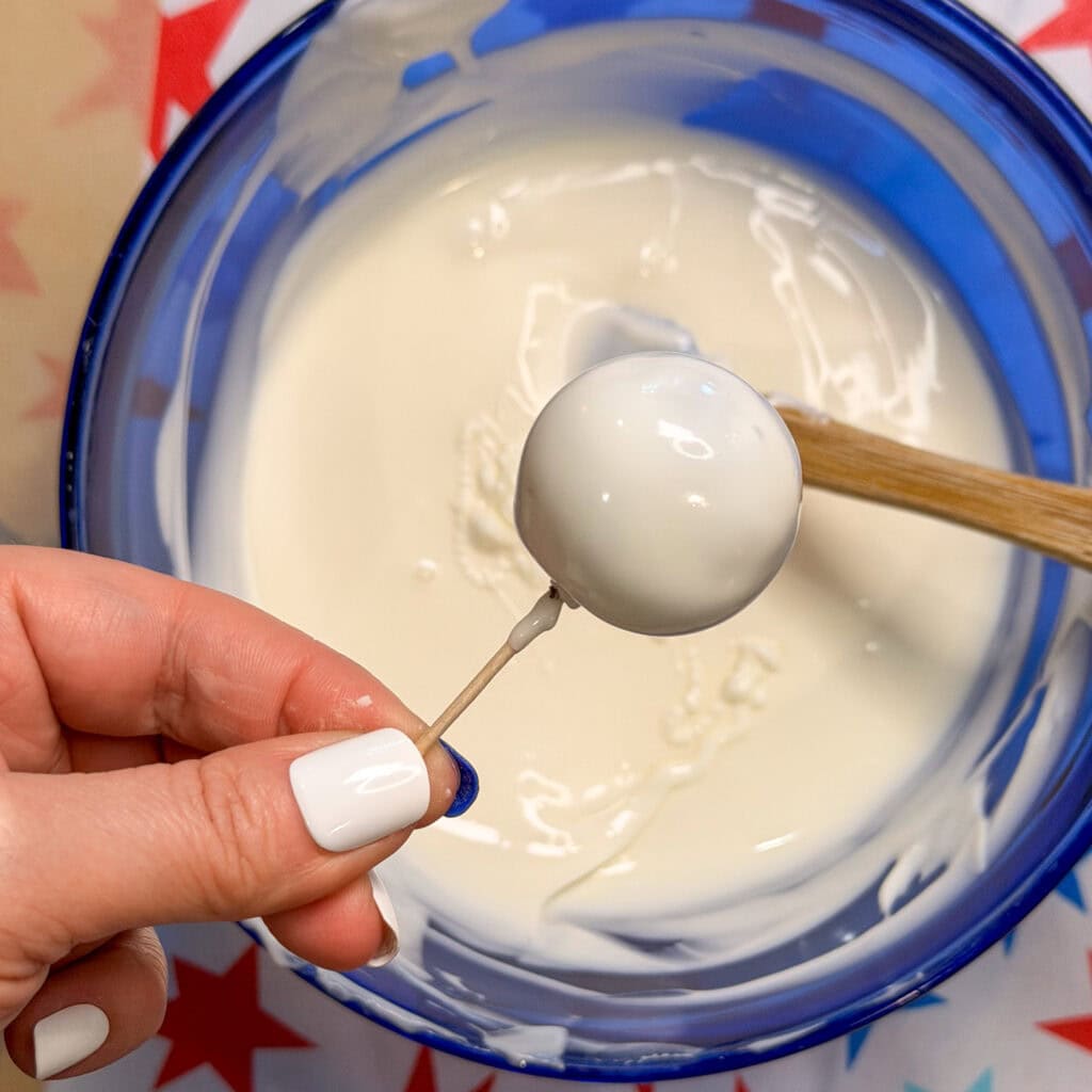 A hand holding a cake pop dipped in white icing above a blue bowl filled with white melted chocolate, similar to how Patriotic Oreo Truffles are prepared. A wooden spoon rests inside the bowl, and the nails are painted white.