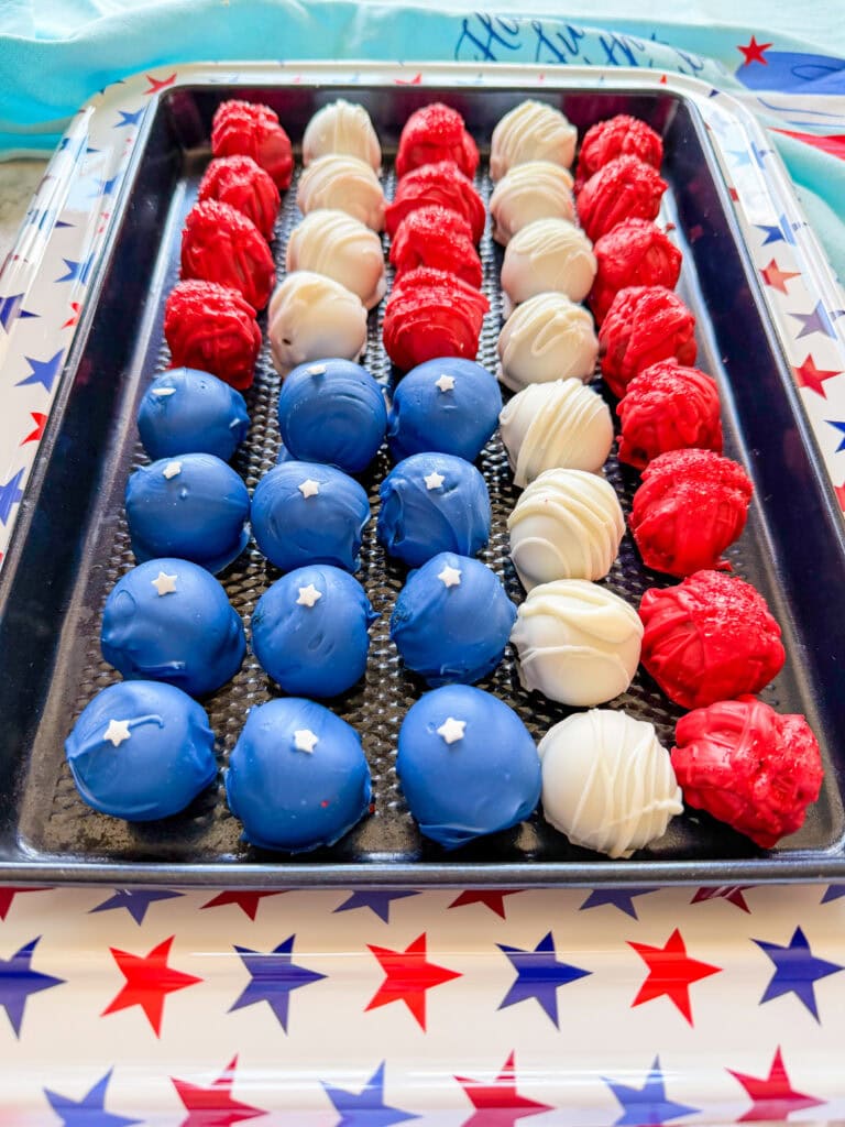 A tray of Patriotic Oreo Truffles arranged in rows of red, white, and blue, decorated with white stars and stripes to form an American flag pattern. The tray and table feature red and blue star designs.