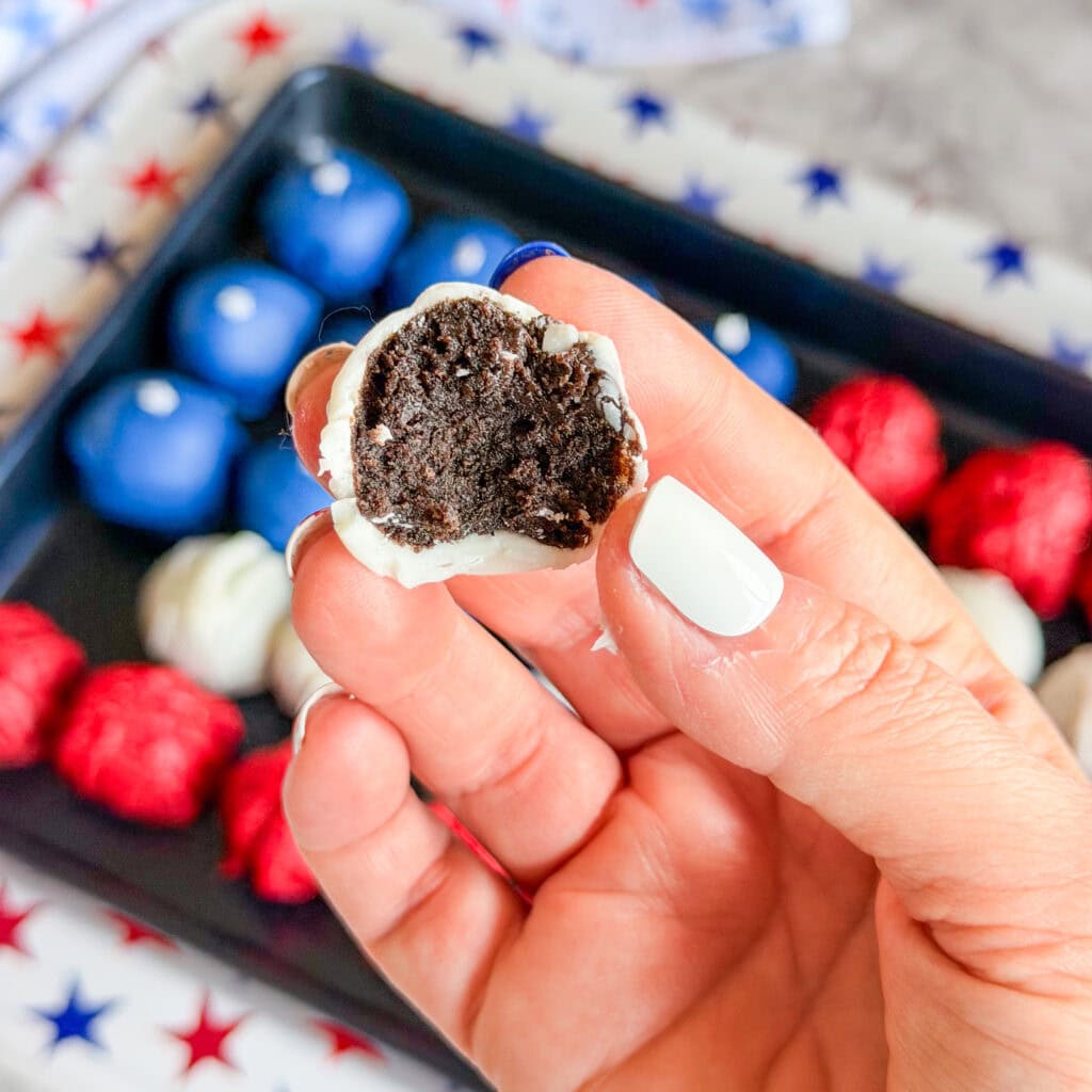 A hand with white-painted nails holds a bitten chocolate truffle coated in white chocolate. In the background, a tray displays Patriotic Oreo Truffles—blue, red, and white—on a star-patterned surface.