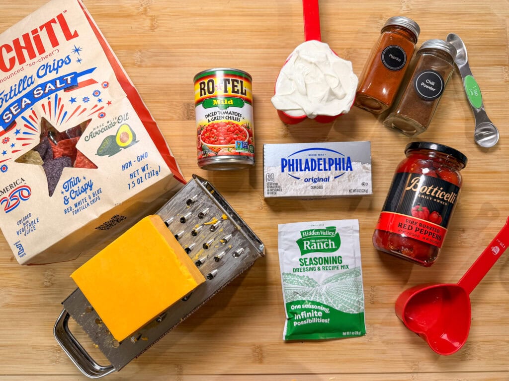 A selection of ingredients on a wooden surface, perfect for making The Best Party Dip—featuring tortilla chips, a can of Rotel, sour cream, cheddar cheese, ranch seasoning, Philadelphia cream cheese, red pepper flakes, chili powder, and measuring spoons.