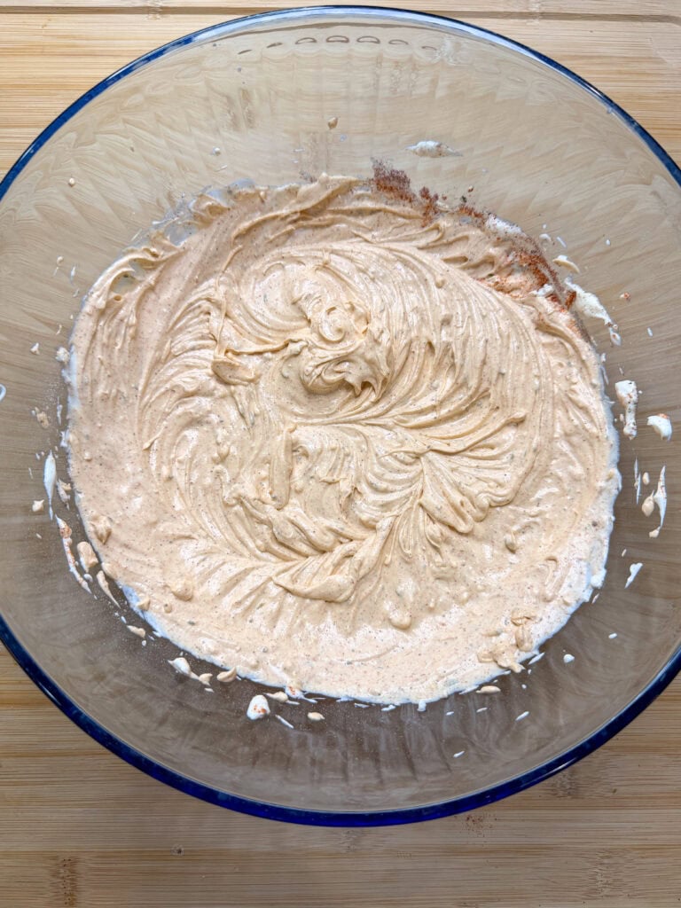 A glass bowl contains The Best Party Dip—a creamy, light brown mixture with visible swirls—sitting on a wooden surface. The dip appears to be partially blended, ready to delight your guests.
