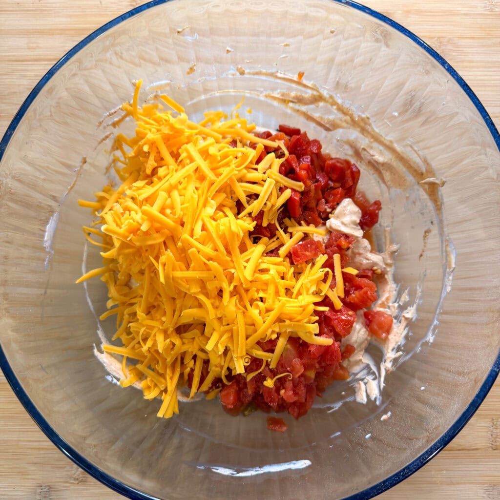 A glass bowl on a wooden surface contains shredded cheddar cheese, diced tomatoes, and chunks of cooked chicken—everything you need to whip up The Best Party Dip, ready to be mixed and enjoyed.