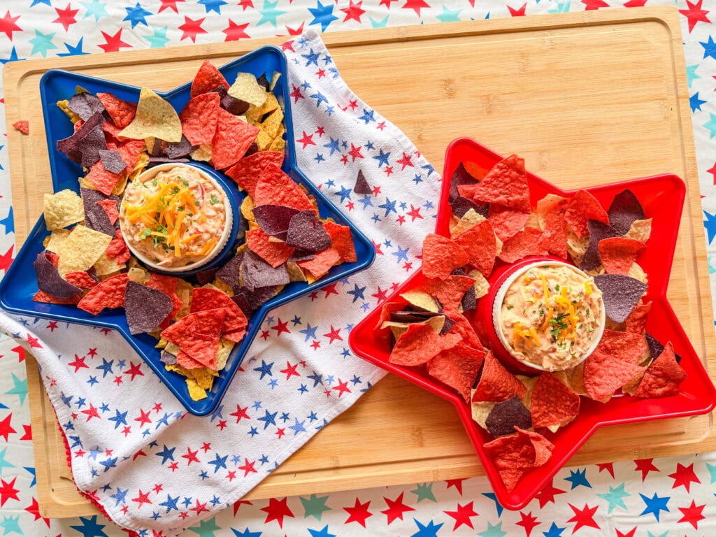 Star-shaped blue and red trays filled with red, blue, and yellow tortilla chips and The Best Party Dip sit on a star-patterned cloth atop a wooden board, creating a festive patriotic display.