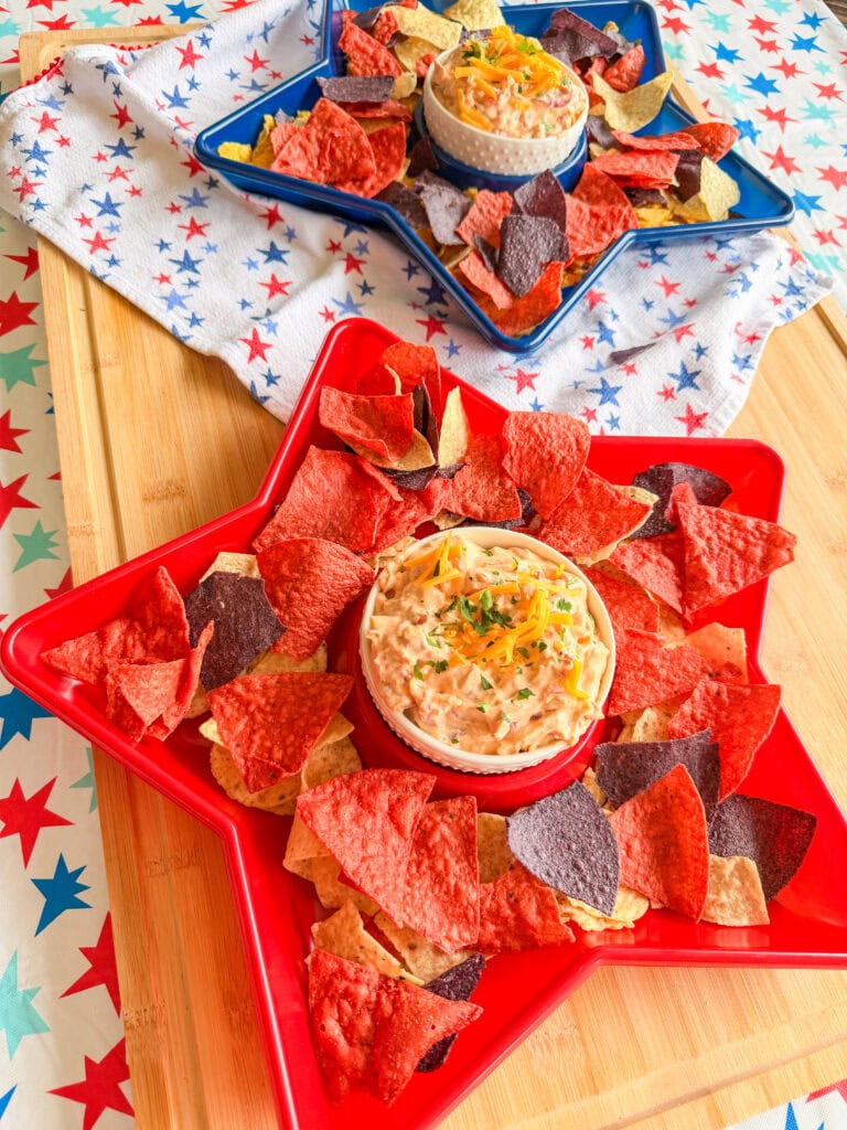 Two star-shaped trays, one red and one blue, are filled with colorful tortilla chips surrounding bowls of The Best Party Dip. Set on a star-patterned cloth and wooden surface, this festive display creates a lively, patriotic theme.