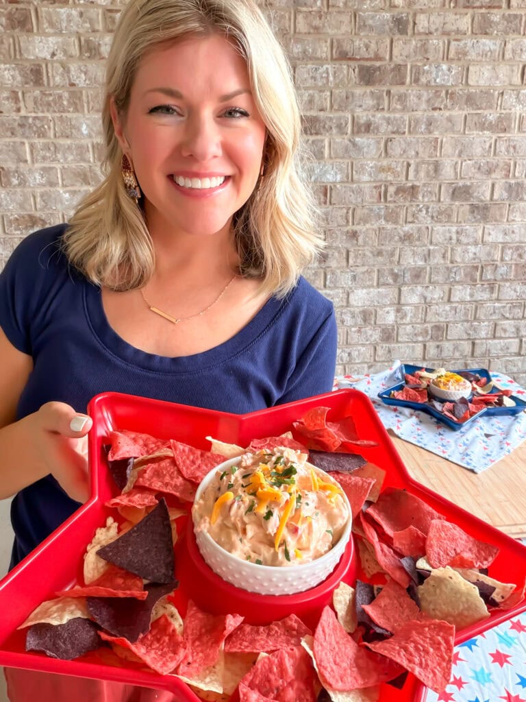 A smiling woman with blonde hair holds a red star-shaped tray with tri-color tortilla chips and a bowl of The Best Party Dip. In the background, another tray with snacks sits on a table against a brick wall.