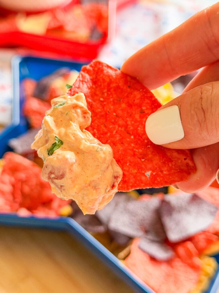 A hand with white nail polish holds a red tortilla chip topped with creamy dip and herbs—perfectly showcasing The Best Party Dip, with more chips and dip in a divided tray in the background.