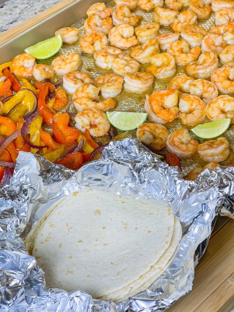 A sheet pan with cooked seasoned shrimp, lime wedges, sautéed bell peppers and onions, and flour tortillas wrapped in foil—ready for assembling shrimp fajitas and perfect for summer recipes.