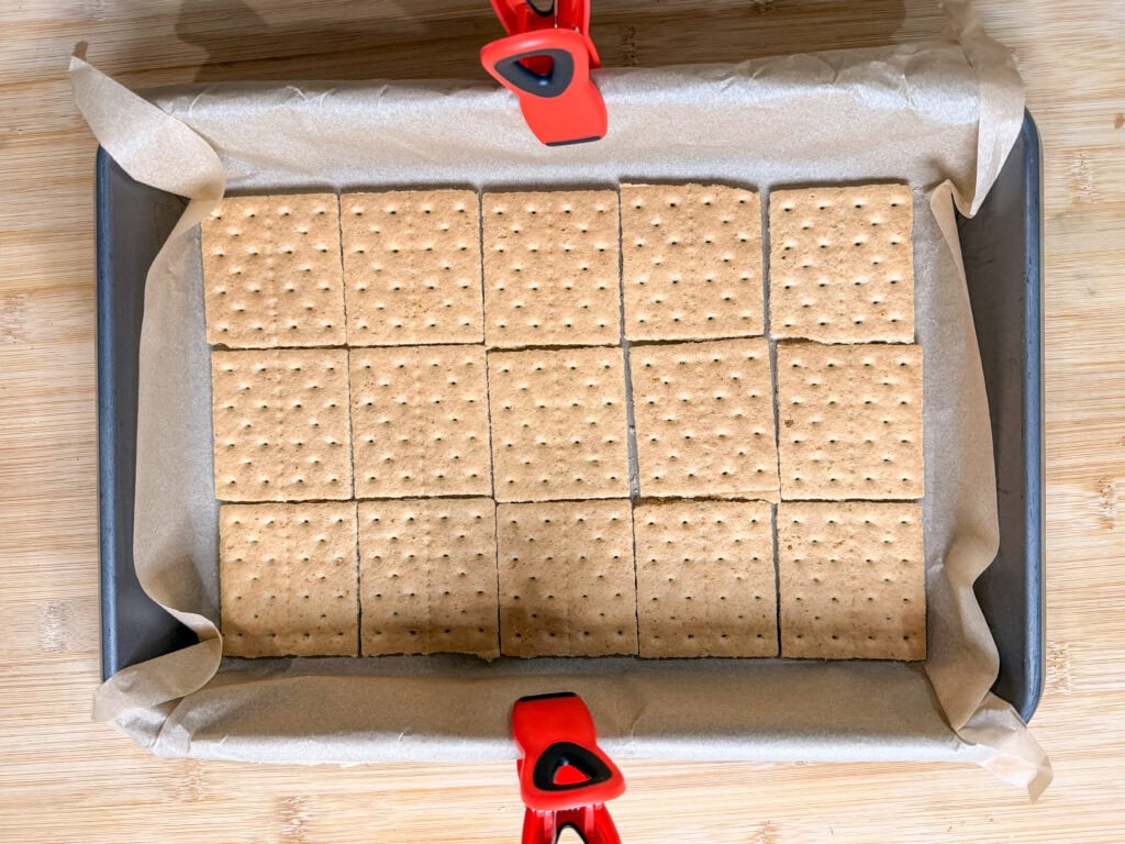 A baking pan lined with parchment paper holds fifteen rectangular graham crackers arranged in a 3x5 grid, ready to become Frozen S'mores, with red clips securing the parchment to the pan on two sides.