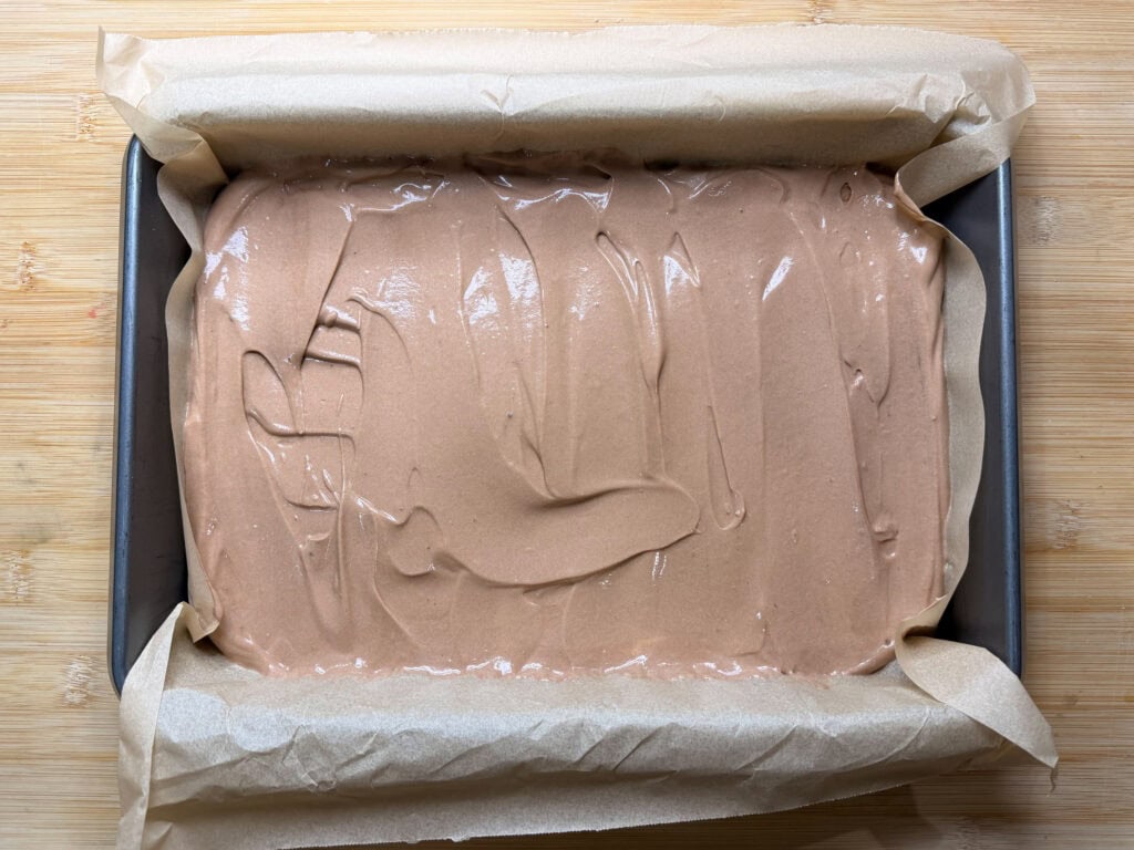 A rectangular baking pan lined with parchment paper is filled with smooth, light brown Frozen S'mores chocolate cake batter, ready to be baked, sitting on a wooden surface.