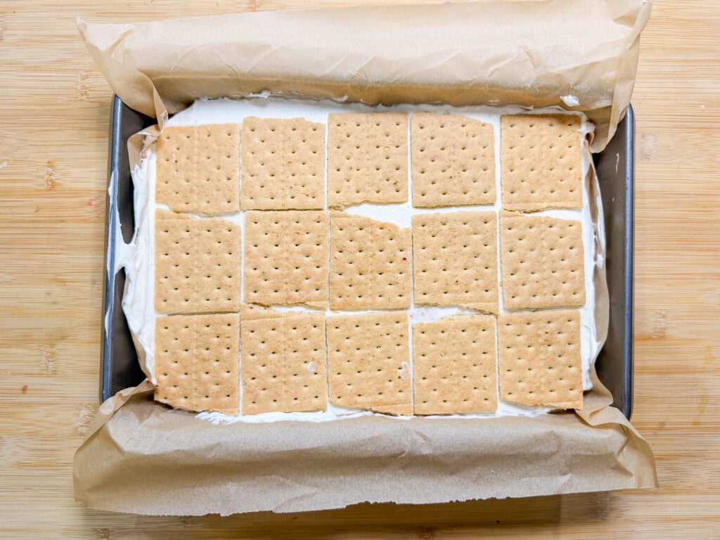 A rectangular baking tray lined with parchment paper contains a layer of cream topped with evenly arranged graham crackers, creating the base for delicious Frozen S'mores, seen from above on a wooden surface.