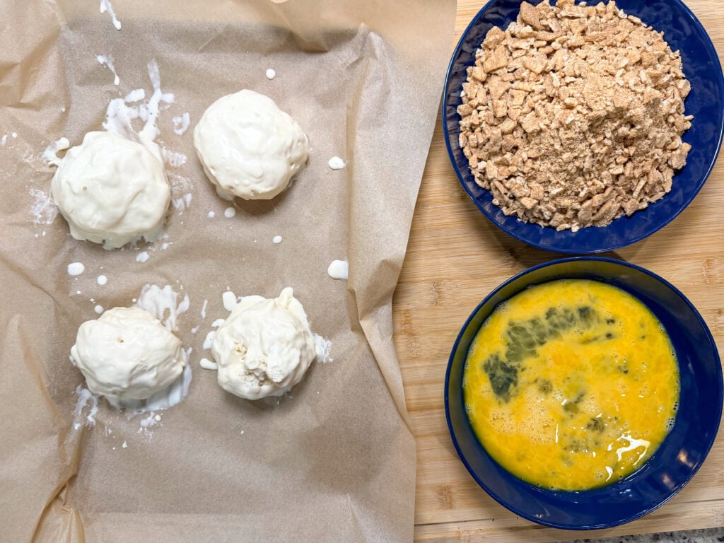Four cream-coated balls on parchment paper beside bowls of crushed crackers and beaten eggs, set on a wooden surface—perfectly prepped for Air Fryer Fried Ice Cream breading or coating.
