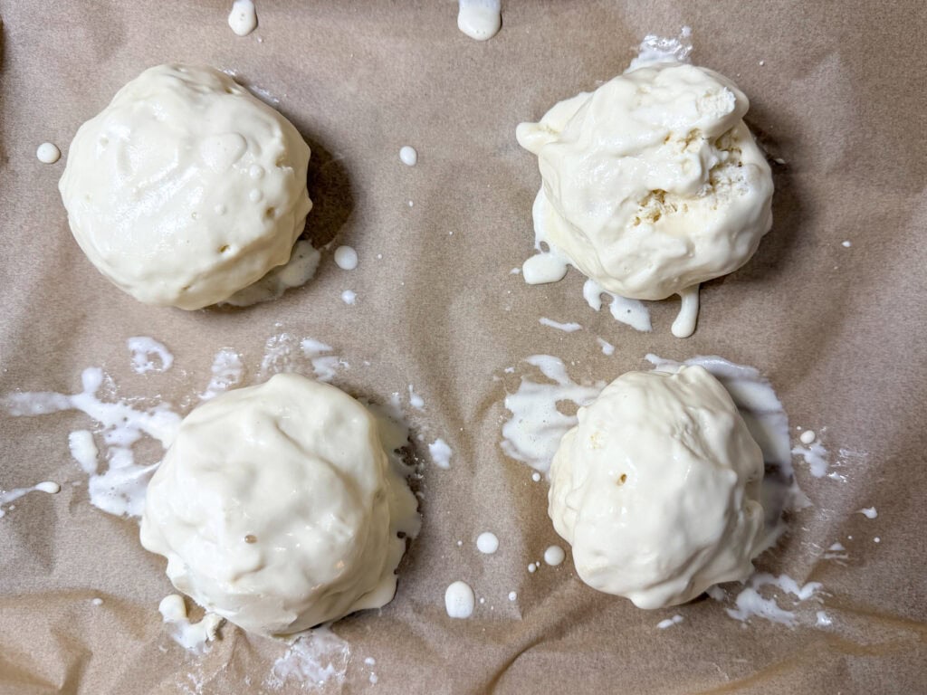 Four unbaked dough balls coated in a white batter are placed on a sheet of brown parchment paper, ready to become Air Fryer Fried Ice Cream, with some batter drips around each dough ball.