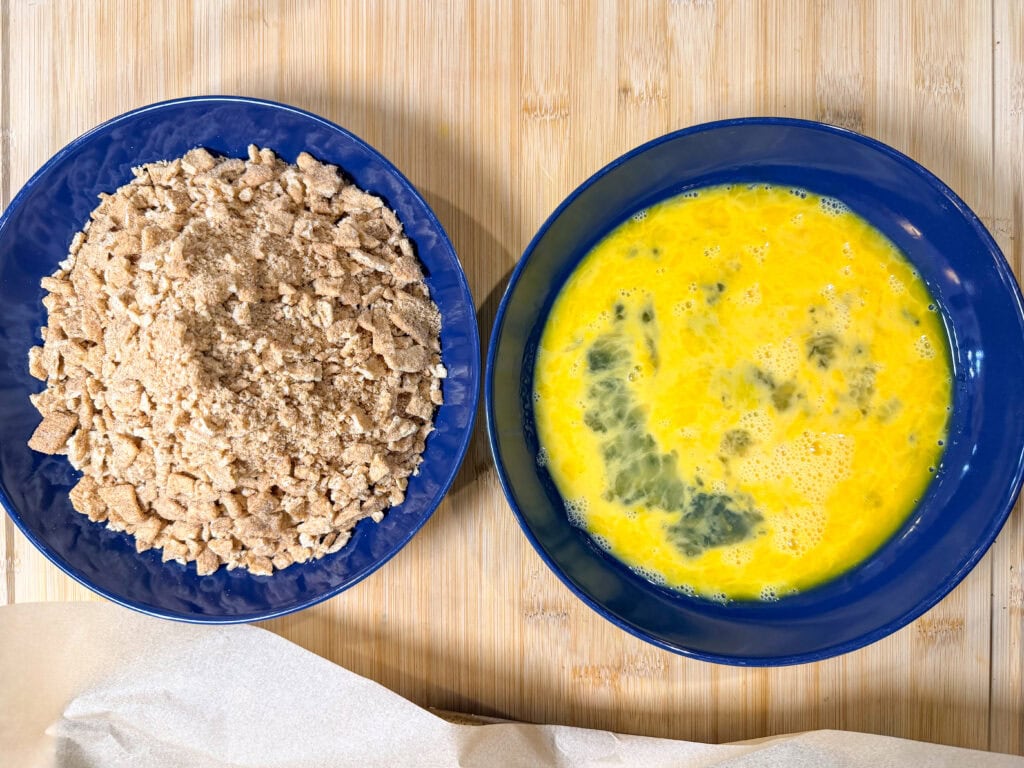 Two blue bowls on a wooden surface: one filled with crumbled bread or breadcrumbs, the other holding whisked eggs with yellow and darker areas—essentials for making Air Fryer Fried Ice Cream. A piece of parchment paper is partially visible below.