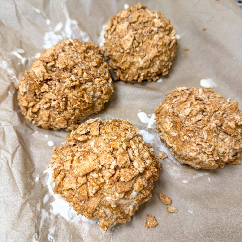 Four round, golden-brown balls of Air Fryer Fried Ice Cream coated in a crumbly cereal crust sit on a sheet of brown parchment paper, with a few spots of white drips around them.