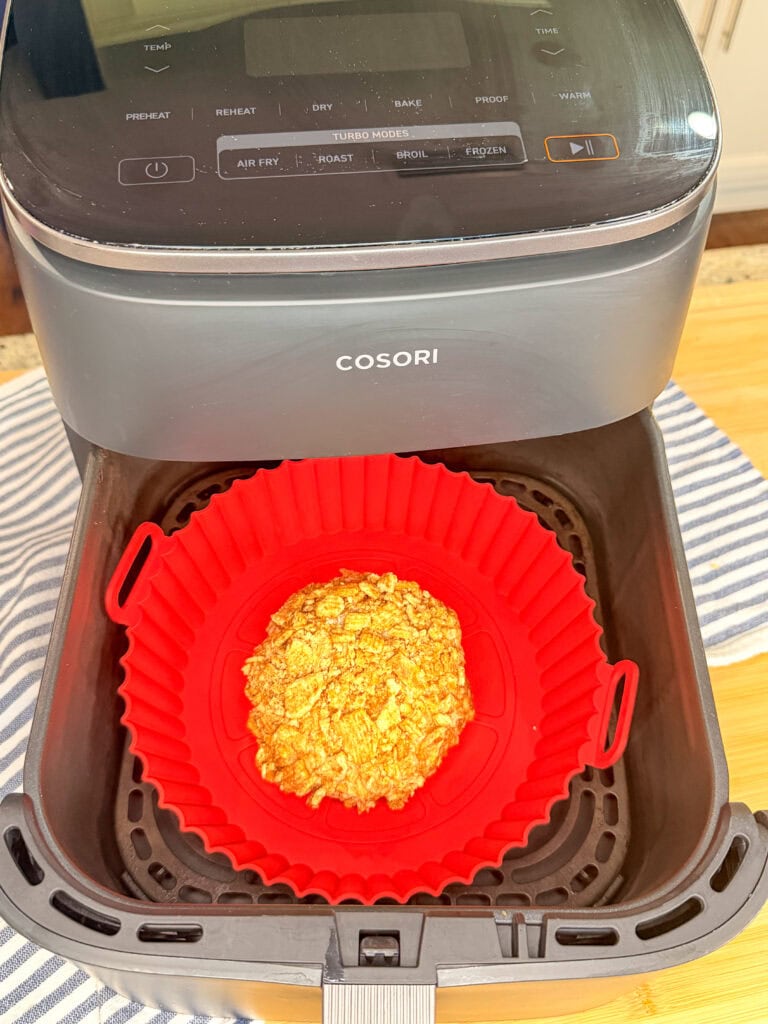 A red silicone liner holding a portion of Air Fryer Fried Ice Cream sits inside the basket of a COSORI air fryer, which is placed on a striped cloth. The air fryer is open and ready for cooking.