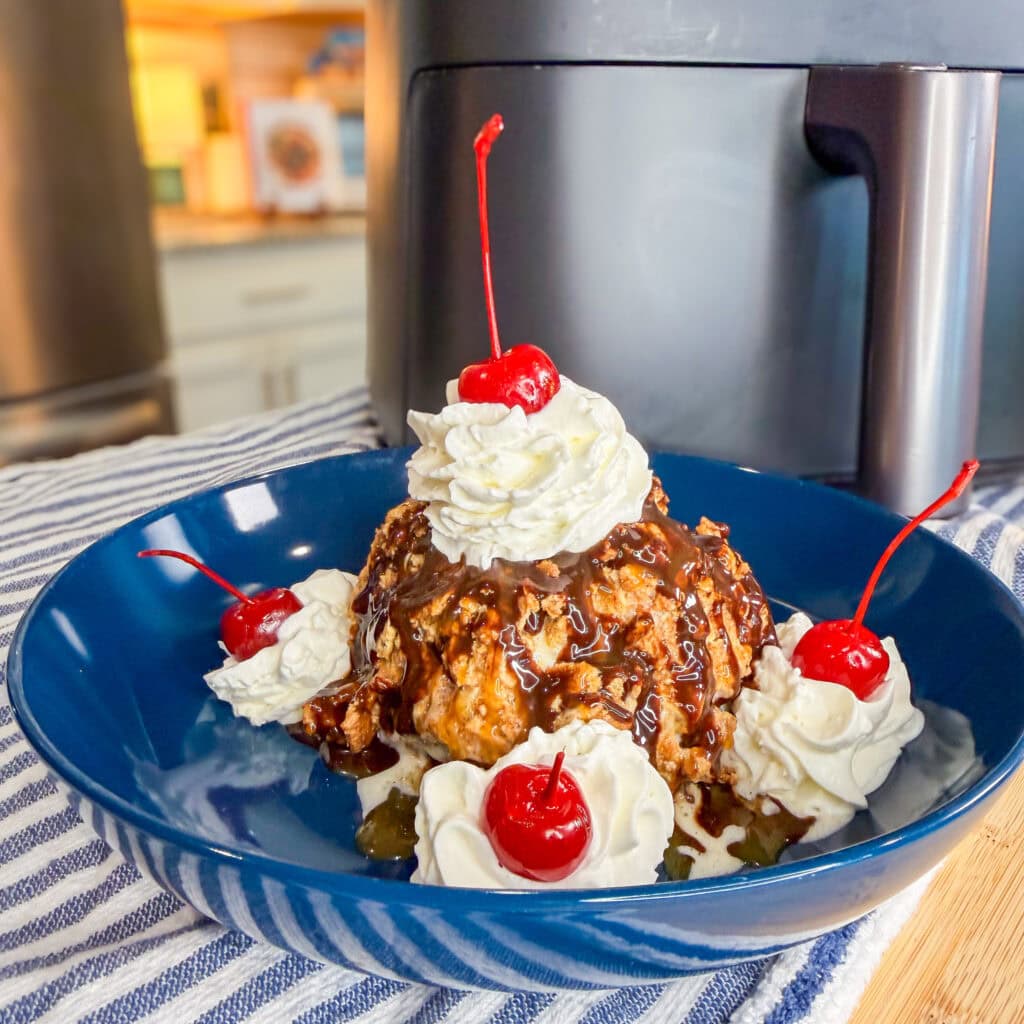 A dessert in a blue bowl showcases Air Fryer Fried Ice Cream topped with chocolate sauce, whipped cream, and cherries, plus extra whipped cream and cherries around it. The bowl sits on a striped cloth near an air fryer in the kitchen.