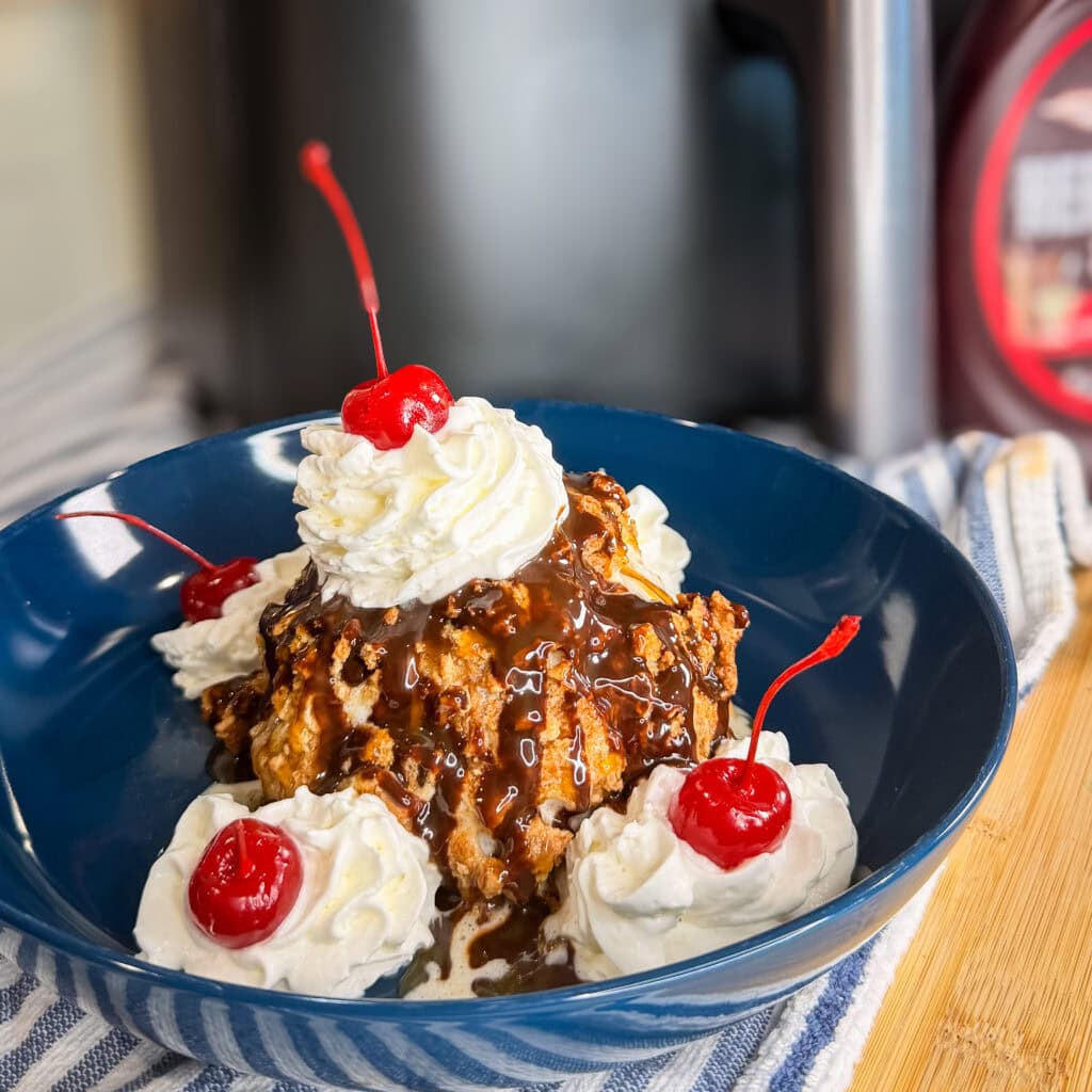A blue bowl holds Air Fryer Fried Ice Cream topped with whipped cream, maraschino cherries, and chocolate sauce, served on a striped cloth with a bottle of chocolate syrup in the background.