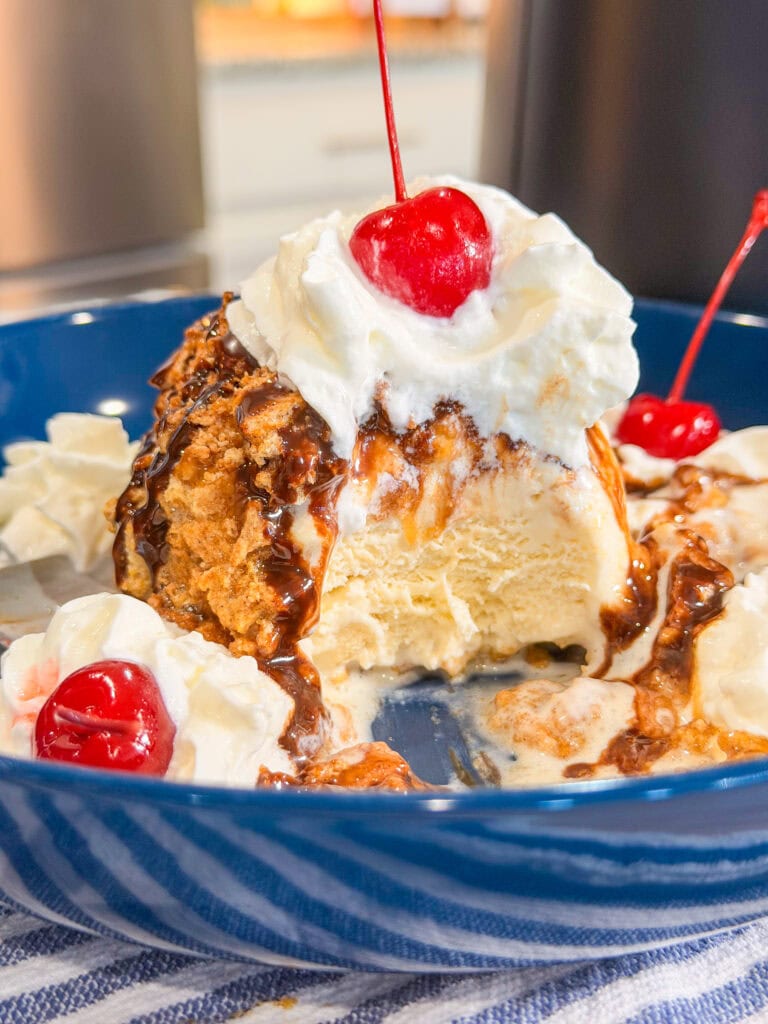 A blue bowl with Air Fryer Fried Ice Cream topped with whipped cream, chocolate sauce, and a cherry. A spoonful has been taken out, revealing vanilla ice cream inside. Whipped cream and cherries decorate the side.