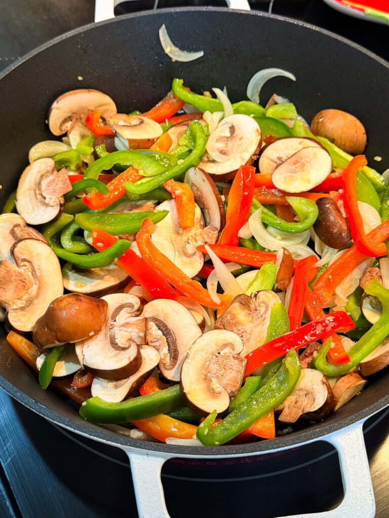 Colorful sliced bell peppers, onions, and mushrooms sautéing in a black pan for a vibrant vegetable medley—the perfect base for a delicious One Pan Cheesesteak Pasta on your stovetop.