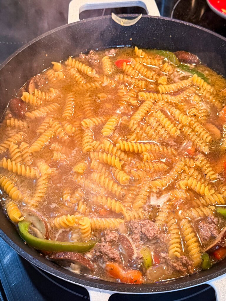 A pot on a stove filled with rotini pasta, ground meat, and vegetables simmering in a savory broth—this One Pan Cheesesteak Pasta fills the kitchen with delicious steam.