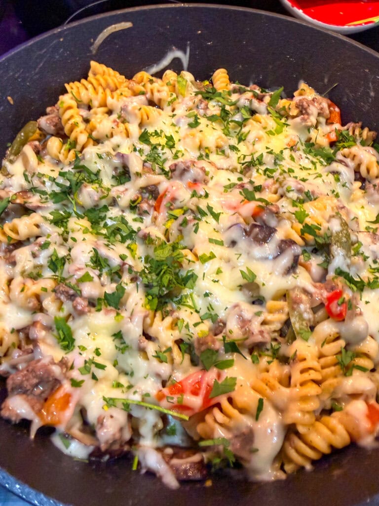 A skillet filled with One Pan Cheesesteak Pasta—rotini, cooked vegetables, ground meat, and melted cheese—all garnished with chopped fresh herbs.