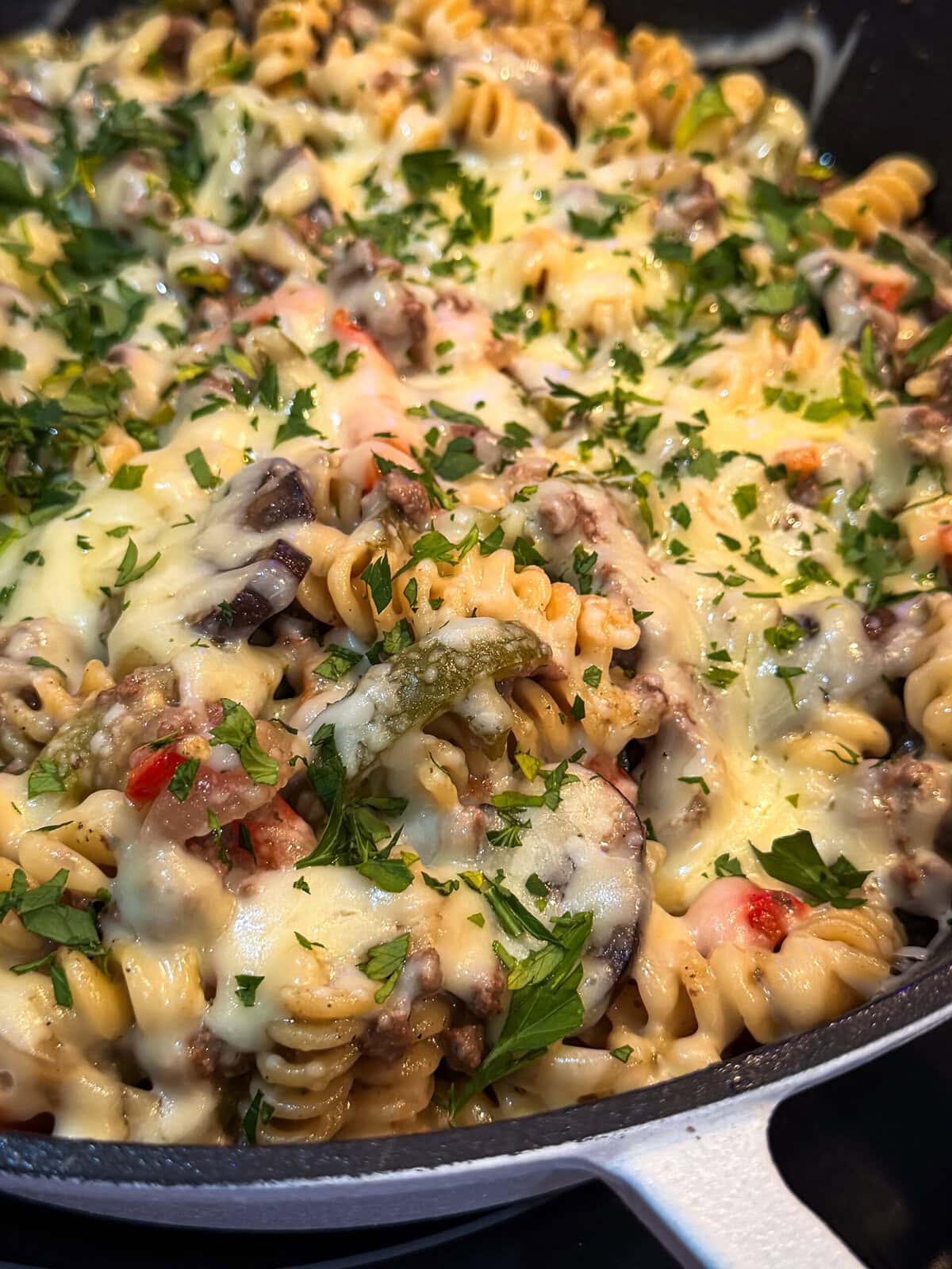 A close-up of One Pan Cheesesteak Pasta, featuring rotini pasta, melted cheese, ground meat, diced tomatoes, mushrooms, and a sprinkle of fresh parsley on top.