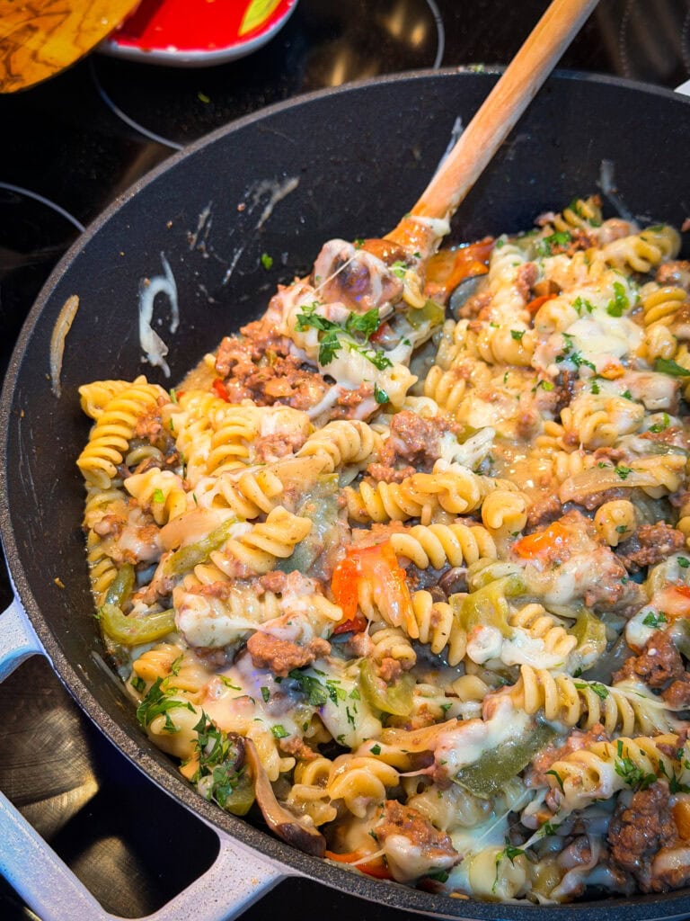 A skillet filled with One Pan Cheesesteak Pasta—rotini, ground meat, bell peppers, herbs, and melted cheese—being stirred with a wooden spoon.