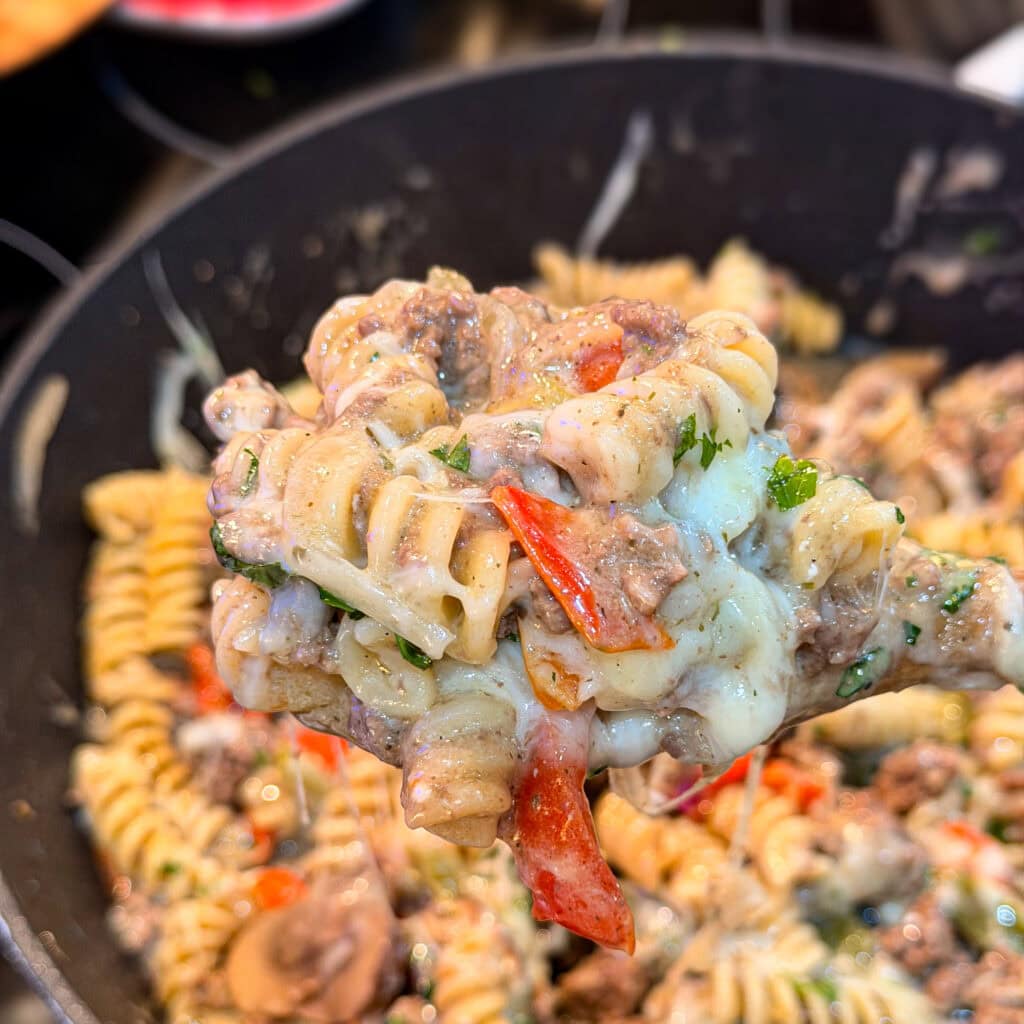 A close-up of a spoonful of cheesy rotini pasta with ground beef, red bell peppers, mushrooms, and herbs—this One Pan Cheesesteak Pasta is held above a pan brimming with the same savory dish.
