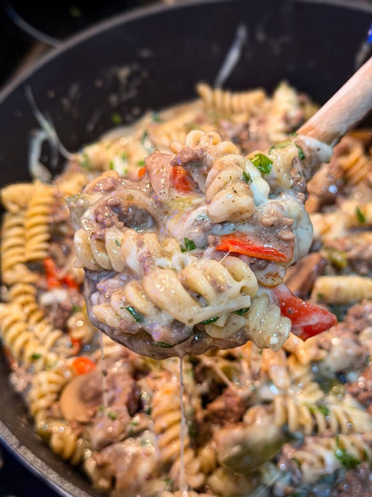 A close-up of a wooden spoon holding cheesy One Pan Cheesesteak Pasta—rotini mixed with ground beef, mushrooms, red bell peppers, and herbs—hovering over a pan filled with the same hearty pasta dish.