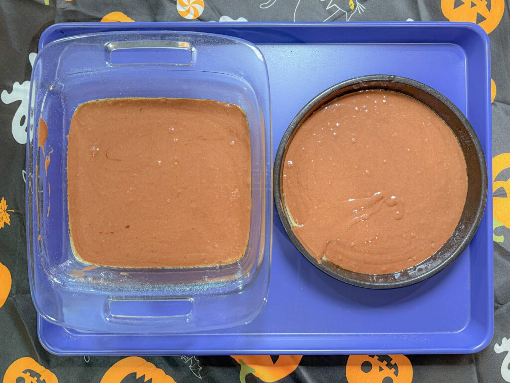 A glass square pan and a round metal pan, both filled with chocolate cake batter, sit on a blue tray—perfect for trying out one of your 10 Easy Halloween Ideas. The background features a Halloween-themed tablecloth with pumpkins and ghosts.