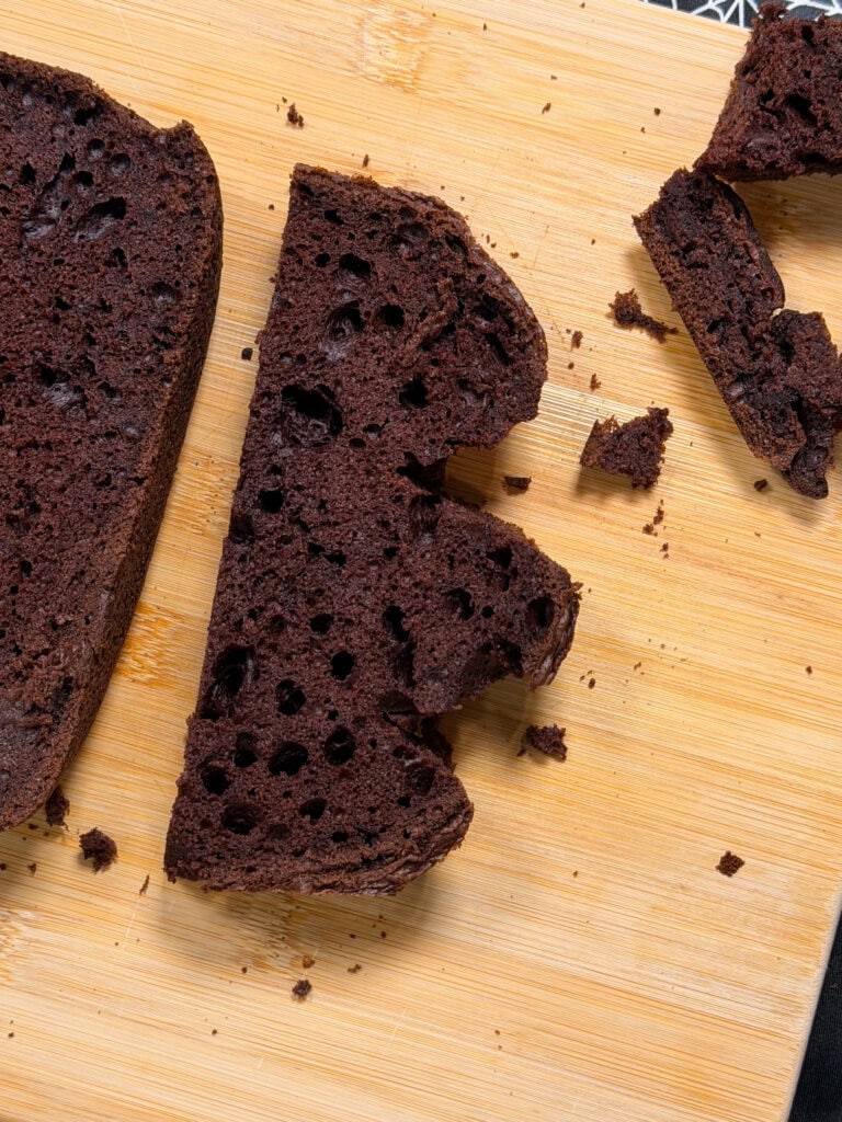 A sliced piece of chocolate bread with large air holes sits on a wooden cutting board, perfect for adding to your spread of 10 Easy Halloween Ideas. The bread is unevenly cut, with a few crumbs scattered around.