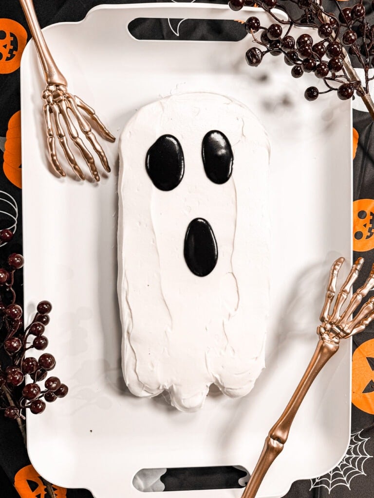 A ghost-shaped cake with white frosting and black candy eyes and mouth sits on a white tray, decorated with skeleton hands, berries, and Halloween-themed designs—perfect as inspiration for your list of 10 Easy Halloween Ideas.