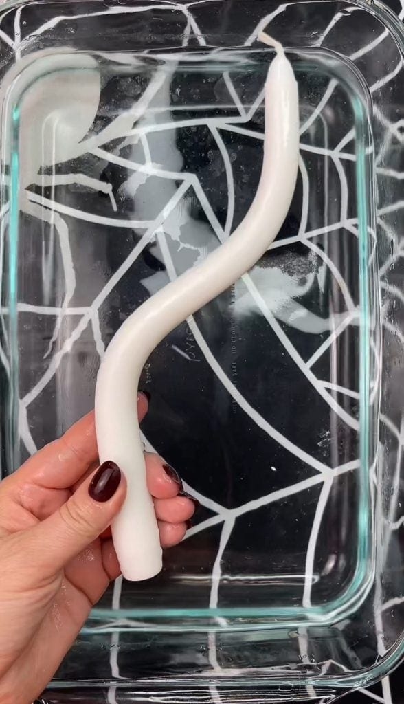 A hand with dark red nails holds a white, curved candle over a clear rectangular dish filled with water—one of 10 Easy Halloween Ideas to set a mysterious mood. The dish sits on a black and white geometric-patterned surface.