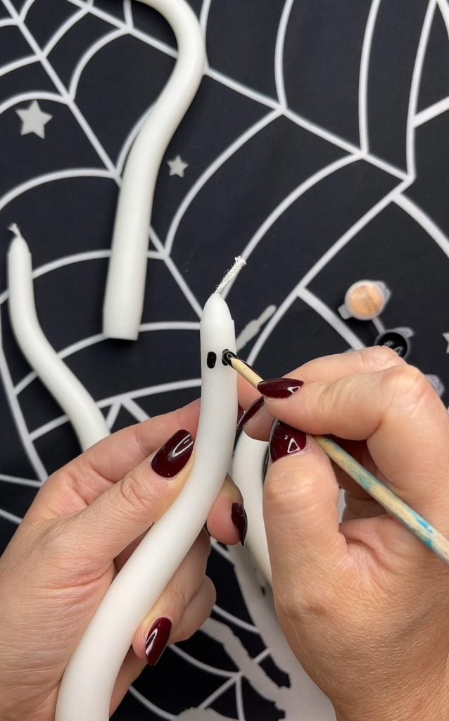 A person paints black eyes on a white, snake-shaped candle, creating a ghost face—a perfect addition to your list of 10 Easy Halloween Ideas. The background has a spiderweb pattern, and other similar candles are visible.