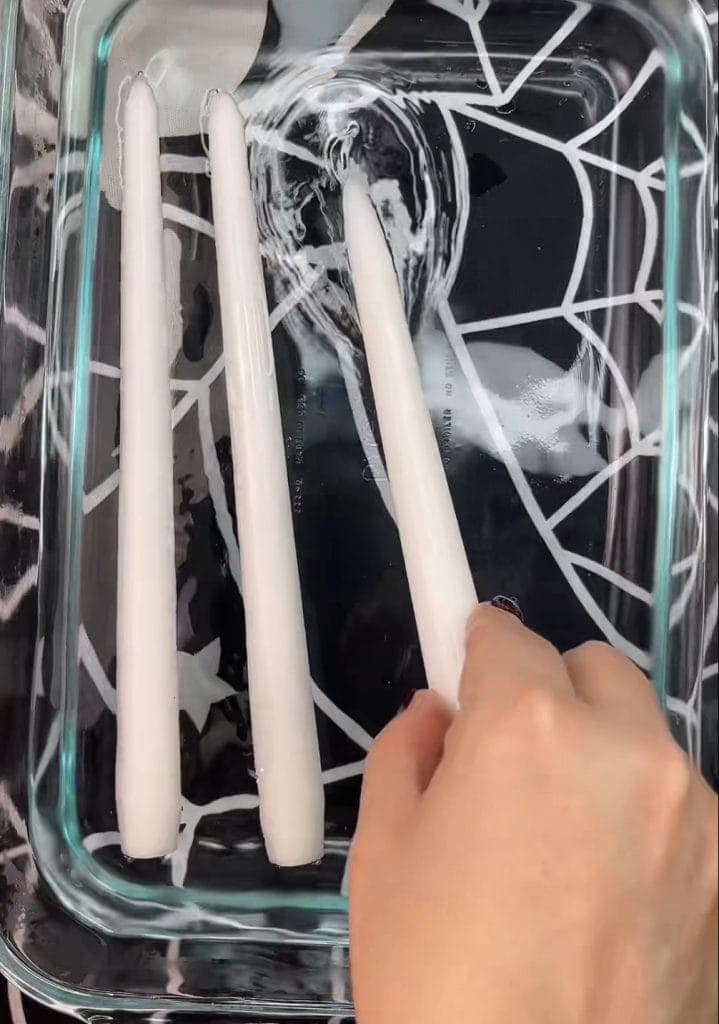 A hand places one of three white taper candles in a glass dish with a black and white spiderweb pattern underneath—one of 10 Easy Halloween Ideas to create a spooky yet elegant centerpiece.