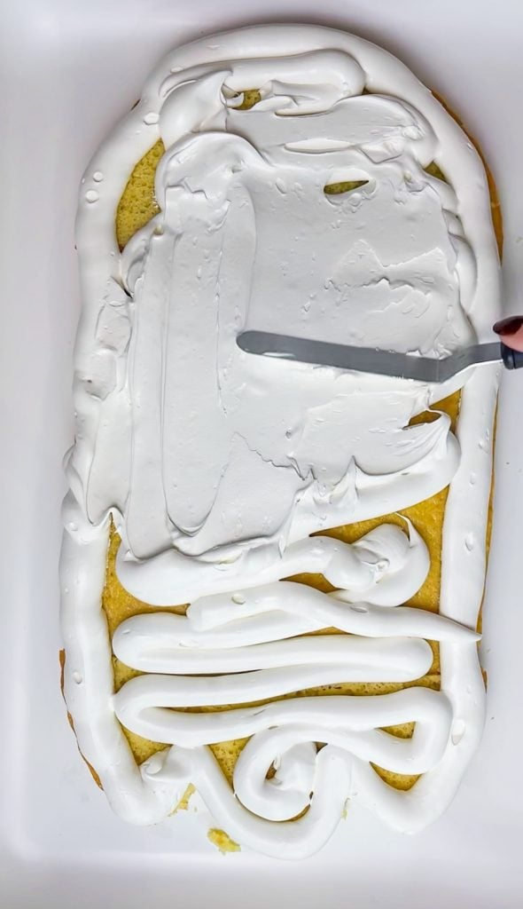 A hand spreads white frosting over a rectangular cake using a metal spatula—one of 10 easy Halloween ideas for festive treats. The cake sits in a white dish and appears partially covered, with visible swirls and lines of frosting.
