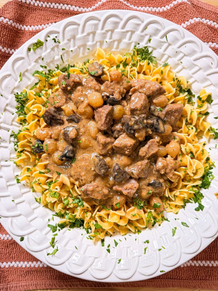 A white plate with Crock Pot Beef and Noodles topped with beef stew, mushrooms, and pearl onions, garnished with chopped parsley, on a textured orange cloth.
