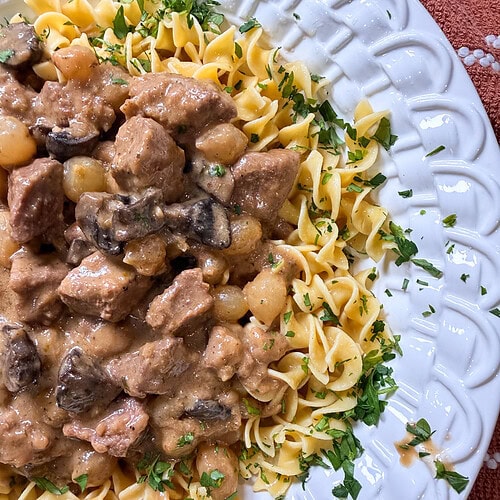 A white plate of egg noodles topped with creamy Crock Pot Beef and Noodles stew, rich with tender beef and savory mushrooms, garnished with chopped fresh parsley.
