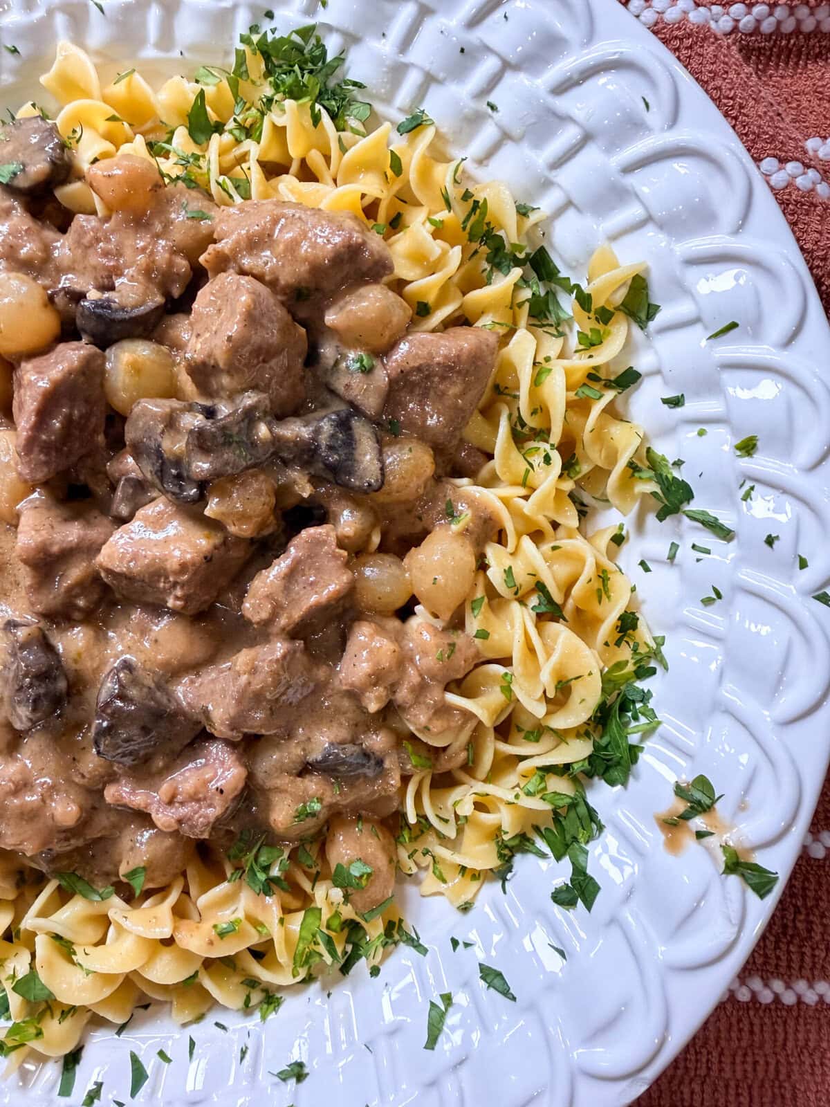A white plate of egg noodles topped with creamy Crock Pot Beef and Noodles stew, rich with tender beef and savory mushrooms, garnished with chopped fresh parsley.