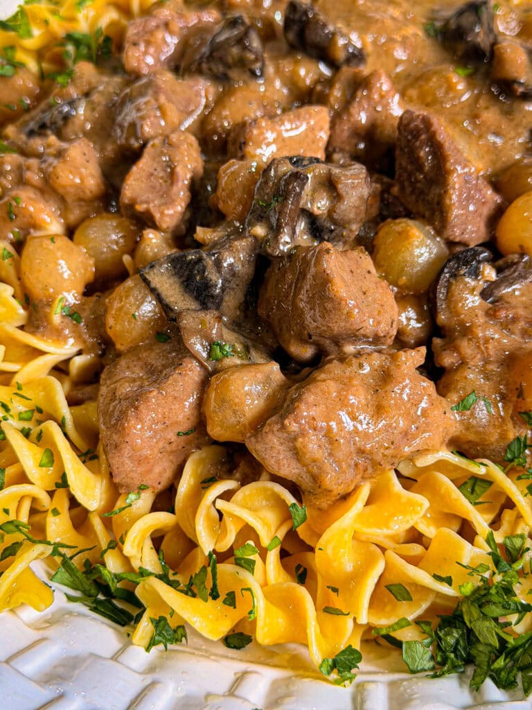 Close-up of Crock Pot Beef and Noodles, featuring tender chunks of beef, mushrooms, and onions in a creamy sauce served over egg noodles and garnished with chopped parsley.