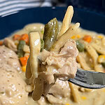 A close-up of a fork holding creamy Crock Pot Chicken and Noodles, with tender chicken, noodles, green beans, peas, and carrots lifted above a bowl filled with the same hearty dish.