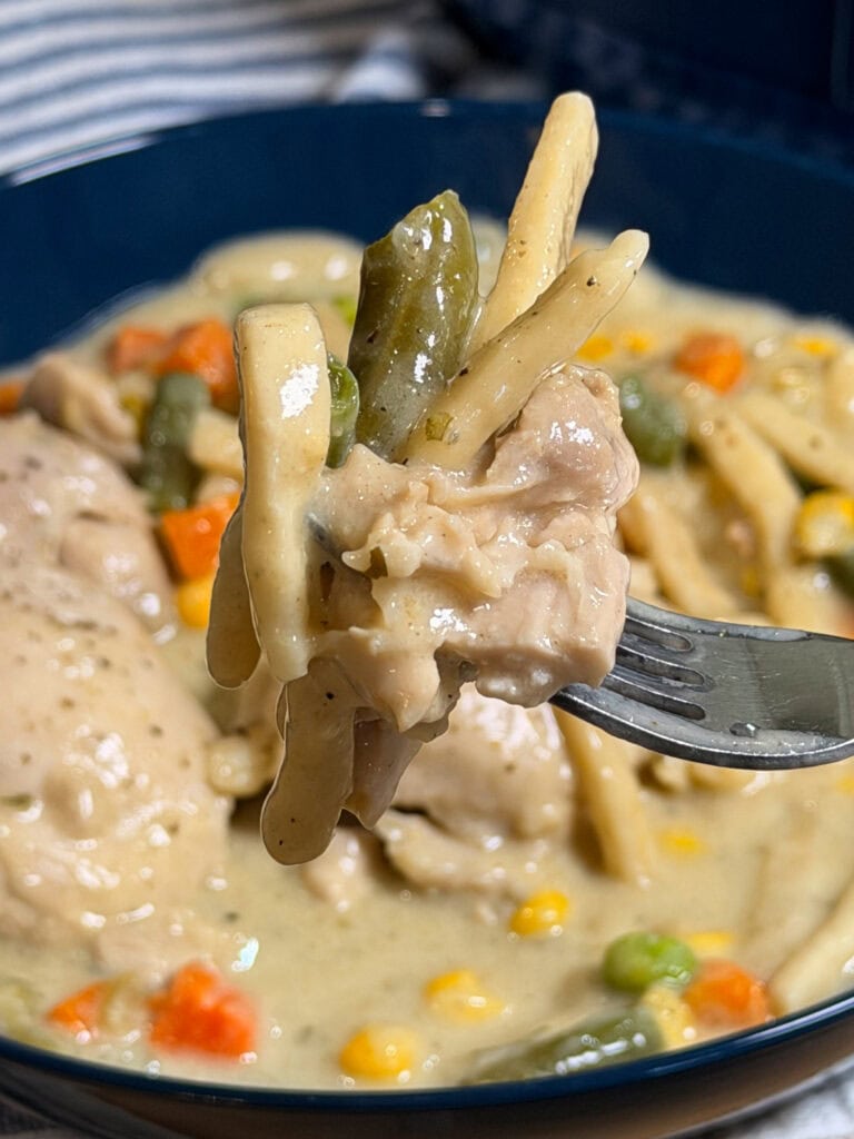 A close-up of a fork holding creamy Crock Pot Chicken and Noodles, with tender chicken, noodles, green beans, peas, and carrots lifted above a bowl filled with the same hearty dish.