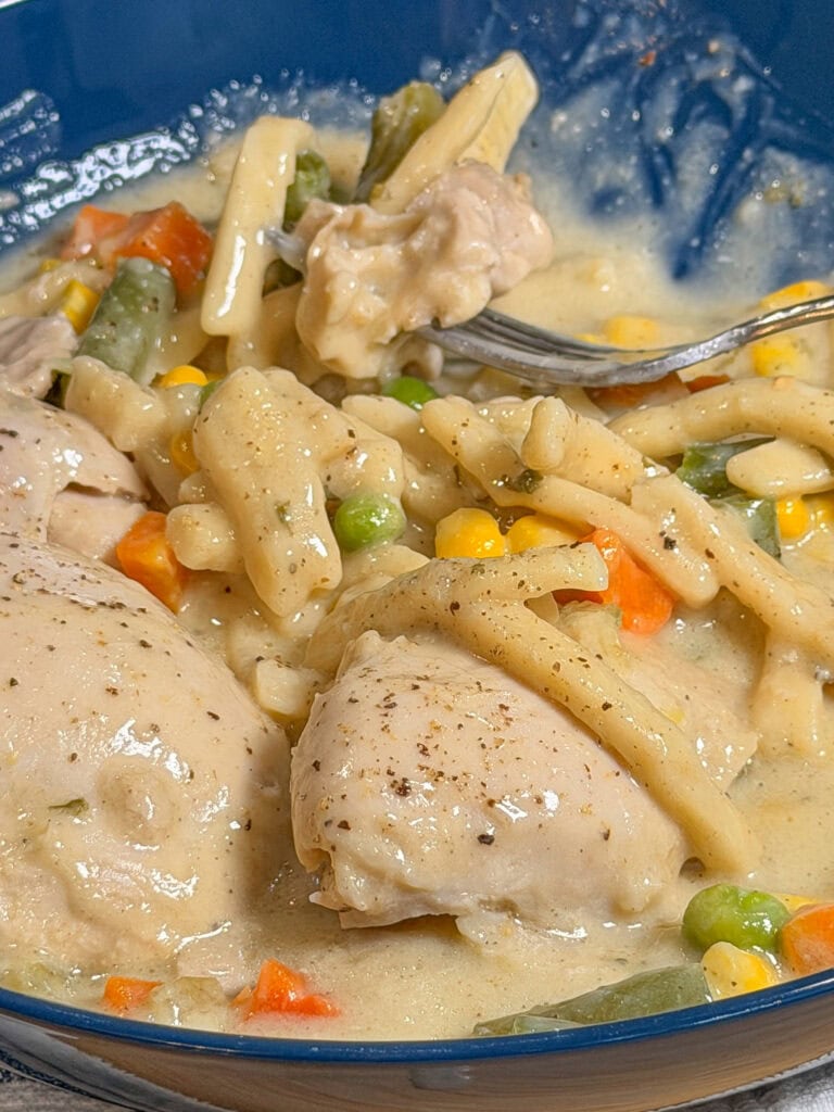 A close-up of a creamy Crock Pot Chicken and Noodles stew in a blue bowl, featuring pieces of chicken, colorful mixed vegetables, and thick noodles, with a fork resting in the food.