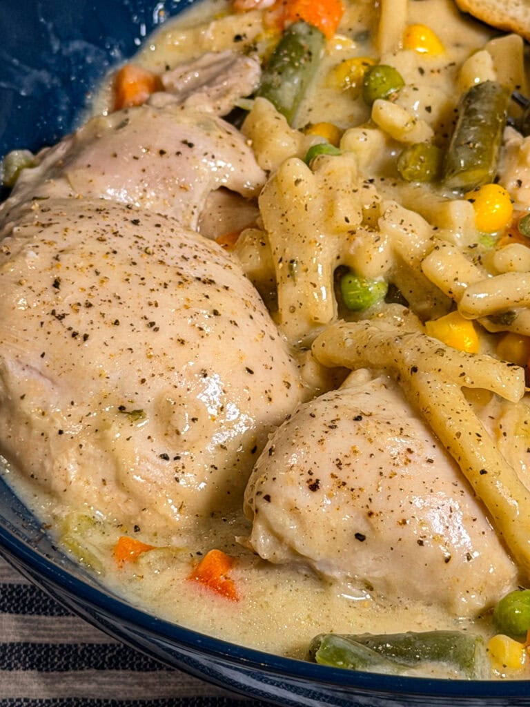 Crock Pot Chicken and Noodles features tender chicken thighs in a creamy sauce with macaroni noodles, mixed vegetables including corn, carrots, peas, and green beans, all served in a blue bowl.