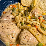 A blue bowl filled with creamy Crock Pot Chicken and Noodles, combined with mixed vegetables like carrots, peas, green beans, and corn, served with a slice of bread.