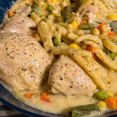 A blue bowl filled with creamy Crock Pot Chicken and Noodles, combined with mixed vegetables like carrots, peas, green beans, and corn, served with a slice of bread.