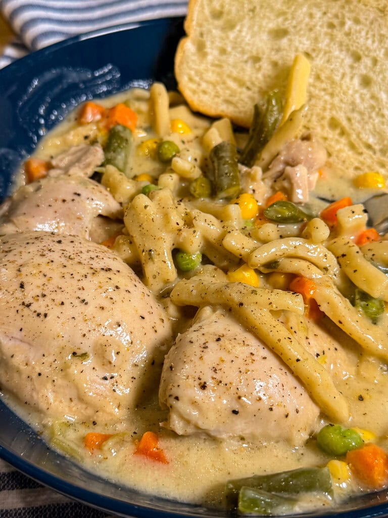 A blue bowl filled with creamy Crock Pot Chicken and Noodles, combined with mixed vegetables like carrots, peas, green beans, and corn, served with a slice of bread.