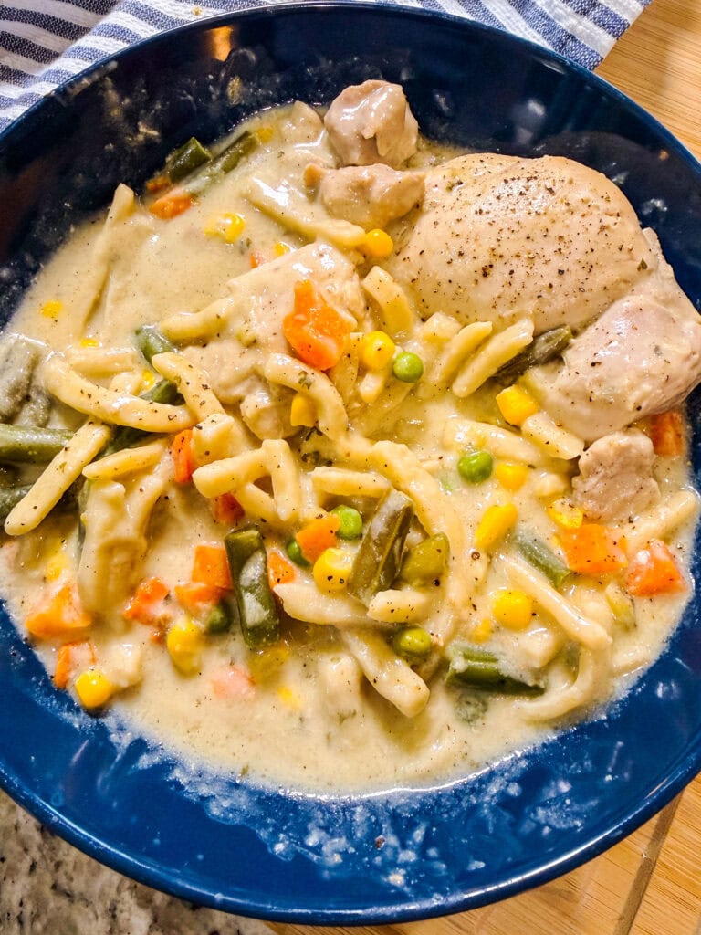 A blue bowl filled with creamy Crock Pot Chicken and Noodles, featuring egg noodles, carrots, green beans, corn, peas, and tender chunks of chicken. The dish appears seasoned and hearty.