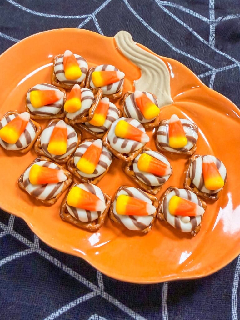Pumpkin-shaped orange plate holding pretzel bites topped with striped chocolate candies and candy corn, placed on a dark blue cloth with a white spiderweb pattern—one of 10 Easy Halloween Ideas for festive party treats.