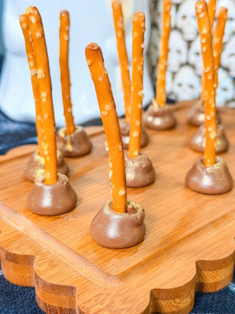 Pretzel sticks inserted into chocolate-covered candies are arranged upright on a wooden serving board, making for one of 10 Easy Halloween Ideas. The candies have a slight golden shimmer, and the background shows blurred decorative objects.